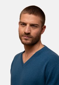 Modrý svetr s výstřihem do V vyrobený z jemné, texturované tkaniny. Design zahrnuje úzký střih a je stylizován s čistým, minimalistickým provedením.