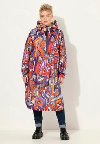 Väriä täynnä oleva pitkä takki, jossa on graffiti-tyylinen kuvio punaisessa, purppurassa ja oranssissa, hupulla ja vetskarilla. Yhdistetty tummiin farkkuihin ja saappaisiin.