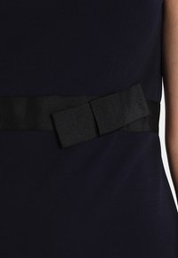 Marindress med ett texturerat svart band i midjan, prytt med en stor rosettdetalj som framhäver kontrasten mellan den släta tyget och det accentuerade metallföremålet.