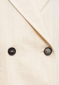 Φωτεινό μπεζ διπλόμπουκωμα σακάκι με υφασμάτινη υφή. Διαθέτει δύο μαύρα κουμπιά με μικρό λογότυπο. Εμφανείς γιακάδες και ραφές.