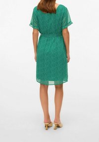 Vestido verde con lunares, con mangas cortas tipo flutter, cintura fruncida y dobladillo a la altura de la rodilla. Tejido ligero y translúcido con una silueta fluida.