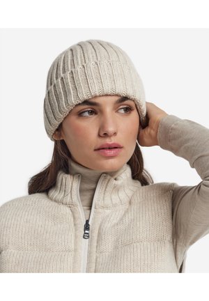 Vrouw draagt een beige gebreide muts en een bijpassende trui met rits, kijkt opzij en raakt met één hand haar hoofd aan.