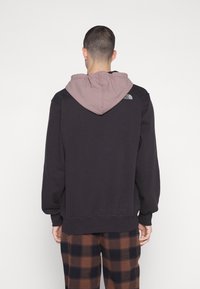 Sudadera negra con capucha rosa claro, con un ajuste relajado y puños acanalados. Logo notable en la parte posterior y tejido texturizado. Pantalones de cuadros marrones.