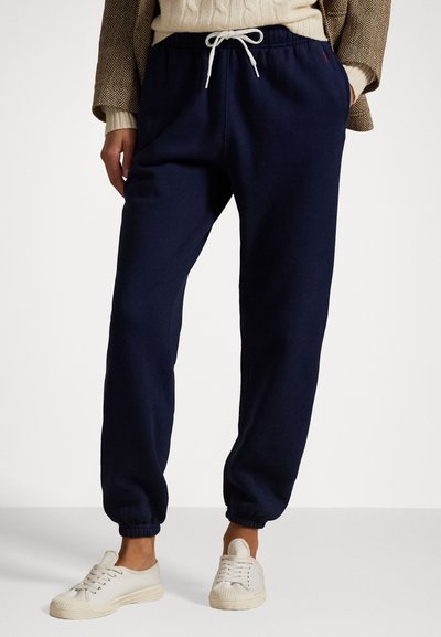 Pantalons de survêtement bleu marine en tissu doux avec des poignets élastiques, dotés d'une taille avec cordon de serrage et de poches latérales, associés à des baskets en toile blanches.