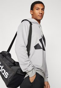 Siva kapuca z malim črnim logotipom Adidas, v kombinaciji s črnim športnim nahrbtnikom z belim napisom. Gladka tekstura, sproščen kroj.