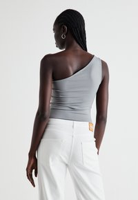 Asymmetrical gray top with a single shoulder strap, sleek fabric, ruching detail, paired with white jeans and a label on the back pocket.