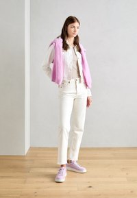 Jeans in denim bianco con orli sfilacciati, una maglietta crema, un maglione rosa a maglia drappeggiato sulle spalle e sneakers lavanda con suole bianche.