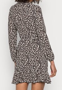 Kobieta w mini sukience z długim rękawem w leoparda, dopasowanej w talii i z falbaną u dołu, stojąca na tle czystego białego tła.
