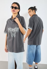 Pelēks polo krekls ar grafisku drukājumu un tauriņa dizainu, kontrastējošām piedurknēm, saskaņots ar džinsu šortiem. Modelis valkā saulesbrilles.