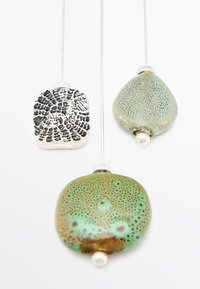 Trois pendentifs sur des chaînes en argent : un pendentif rectangulaire texturé noir, un pendentif en forme de goutte vert avec des éclats, et un pendentif rond vert-brun.