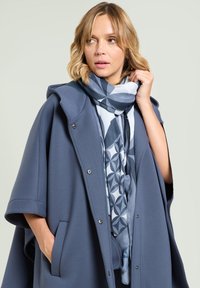 Capa azul oscuro con capucha, hecha de tela suave, con bolsillos laterales y cierres a presión. Se lleva alrededor del cuello una bufanda ligera con estampado en tonos azules.