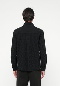 Egy férfi áll, háttal a kamerának, fekete hosszúujjú inget viselve, amelyen diszkrét kör mintázat látható, valamint fekete nadrágot; a háttér egyszínű.