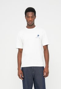 Λευκό βαμβακερό t-shirt με στρογγυλή λαιμόκοψη και κοντά μανίκια, με μπλε λουλουδάτο λογότυπο στο αριστερό στήθος. Συνδυασμένο με τζιν παντελόνι.