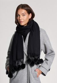 Czarny szalik wykonany z miękkiego materiału, z teksturowanymi frędzlami i okrągłymi, czarnymi pomponami na końcach. Nosi się go na jasnoszarym płaszczu.