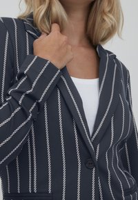 Femme portant un blazer bleu marine à rayures verticales blanches sur un haut blanc, tenant le col de la main droite.