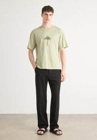 Lys grøn bomulds t-shirt med et palme-træ grafik, parret med sorte bukser og sorte sandaler. Simpelt design med korte ærmer.
