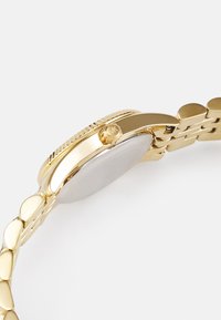 Goldfarbene Uhr mit einem Armband aus Metallgliedern. Sie verfügt über ein Gehäuse aus Edelstahl und eine glatte Lünette, mit einer sichtbaren Krone, die mit "MK" graviert ist.