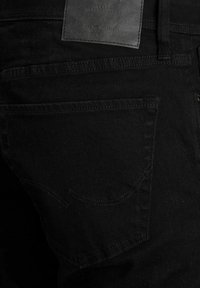 Pantalones vaqueros de mezclilla negra con una textura suave, que presentan costuras en los bolsillos traseros y una etiqueta de cuero en la cintura. Presentan elementos de diseño sutiles.