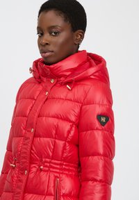 Chaqueta acolchada roja con acabado brillante, que incluye capucha, cordón elástico y detalles en tono dorado. Bolsillos laterales y logo en la manga.