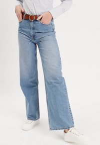 Jean taille haute bleu clair avec un design à jambes évasées, présentant un style classique à cinq poches et une ceinture en cuir marron avec une boucle ronde.