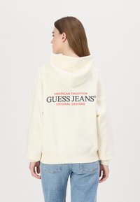 Kremkrāsas kapuce ar lielu drukas attēlu mugurpusē, uzraksts "GUESS JEANS" pelēkā un sarkanā krāsā. Iekļauj tuvējās saites kapuci un brīvu piegriezumu.