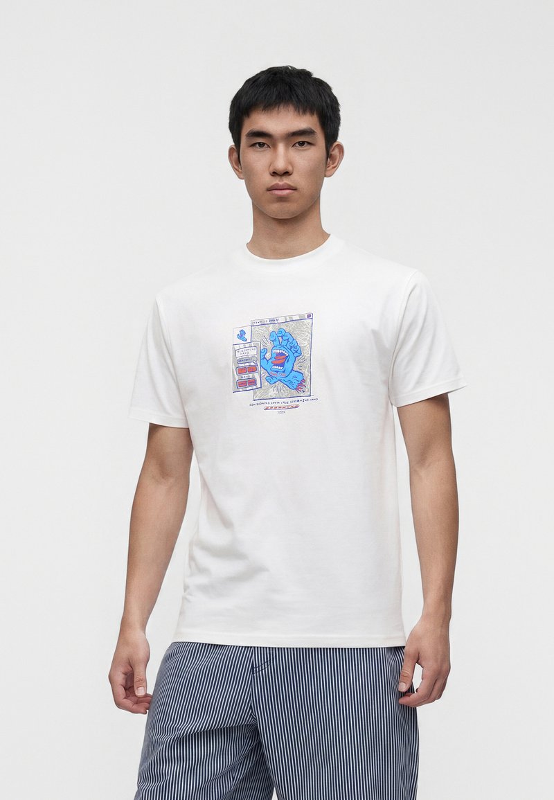 Νέος άντρας που φοράει λευκό T-shirt με μπλε γραφικό μιας ουρλιάζουσας χειρός μέσα σε πλαίσιο παραθύρου, συνδυασμένο με μπλε και λευκές ριγέ παντελόνες.