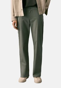 Green, straight-leg pants with a smooth texture, featuring a front button closure and side pockets, worn with beige shoes and a light jacket.