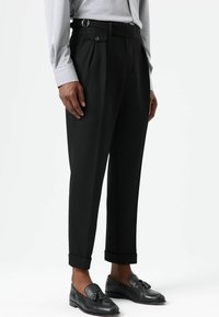 Zwarte pantalons met een getailleerde pasvorm, een platte voorkant, zijzakken en omgeslagen boorden, gecombineerd met zwarte loafers. Soepele textuur met een subtiele glans.