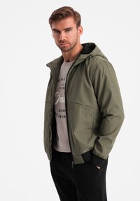 Giacca leggera verde oliva con cappuccio, chiusura con zip e polsini elasticizzati. Indossata sopra una felpa grigia stampata, abbinata a pantaloni neri.