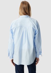 Jasnoniebieska koszula wykonana z bawełny ma długie rękawy, plisę z tyłu i zaokrąglony dół. Zawiera mankiety zapinane na guziki oraz klasyczny kołnierzyk.
