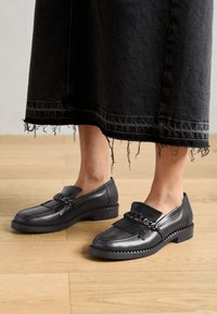 Juodos odinės loafer tipo bateliai su pakraščiu ir grandinėlės detalėmis, su tekstūruota padu ir žemu blokuotu kulnu, dėvimi su nusidėvėjusia džinsine sijonu.