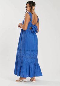 Vestido maxi azul con tirantes con volantes, espalda descubierta y detalle de lazo. Presenta un diseño bordado y un dobladillo en capas con volante. Textura de tela suave.