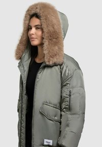 Giacca verde oliva imbottita con cappuccio in pelliccia sintetica marrone staccabile. Presenta tasche con zip e una patch con logo all'orlo inferiore.