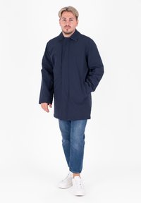 Giacca esterna blu navy con chiusura a bottoni, due tasche laterali e colletto, abbinata a jeans blu chiaro e sneakers bianche.