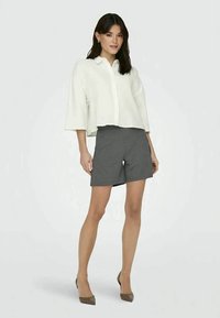 Witte, korte mouwen, geknoopte blouse en grijze op maat gemaakte korte broek. Model draagt beige hoge hakken. Gladde textuur, ontspannen pasvorm.