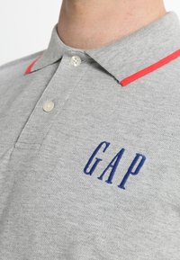 Polo gris avec accents de col rouge, affichant un logo "GAP" brodé en bleu, une patte de boutonnage à deux boutons et un tissu texturé.