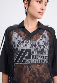 Czarna koszulka Adidas Originals z koronkowym wykończeniem, dekoltem w serek oraz kontrastowymi białymi paskami na rękawach. Na przodzie wyróżniające się logo graficzne.