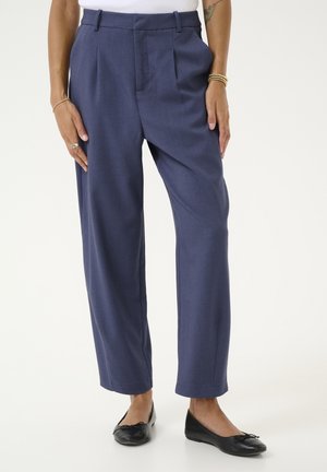 Frau trägt hochtaillierte, marineblaue, zulaufende Hose mit Bundfalten, kombiniert mit schwarzen Ballerinas und einem weißen Oberteil.