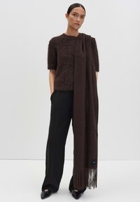 Bruine wollen sjaal met lange franjes, over een kortomarm fuzzy bruin top gedrapeerd, gecombineerd met hoge zwarte broeken. Schone achtergrond.