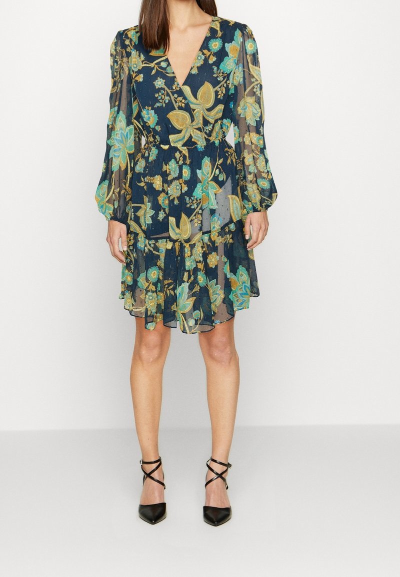 Květované šaty s dlouhými průsvitnými rukávy, dekoltem do V a vrstvenou sukní v tmavě modré barvě se zelenými a žlutými květinovými vzory. Černé lodičky s špičatou špičkou.