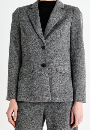 Grå tekstureret to-knaps blazer med klaplommer iført over sort top, parret med matchende grå teksturerede bukser.