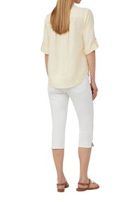 Chemise jaune clair à manches longues avec poignets retroussés, empiècement au dos et ourlet arrondi ; associée à un pantalon Capri blanc et des sandales marron.