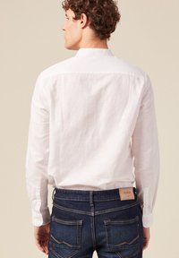 Camicia di lino bianca con maniche lunghe, colletto alla cinese e orlo arrotondato, abbinata a jeans in denim blu scuro con un'etichetta in pelle sulla vita.