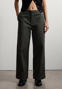 Pantaloni in cotone verde scuro a gamba larga con una texture liscia, dotati di una vita strutturata e orli arrotolati, abbinati a mocassini neri.