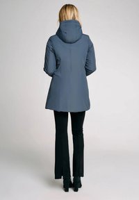 Cappotto isolato grigio scuro con cappuccio, texture liscia e silhouette leggermente svasata, abbinato a pantaloni neri svasati e calzature con tacco.