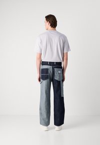 Denim cargo pants with mixed blue shades, featuring multiple pockets and a relaxed fit. Worn with a light gray T-shirt and white sneakers.