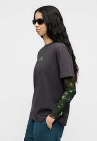 Άτομο με μακριά μαύρα κυματιστά μαλλιά, φοράει μαύρα γυαλιά ηλίου, σκούρο γκρι μπλουζάκι πάνω από πράσινη μπλούζα με μοτίβο μακριάς μανίκας και μπλε παντελόνι, με το ένα χέρι στην τσέπη.