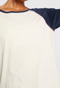 Kortärmad topp med en krämfärgad kropp och marinblå ärmar samt en kontrasterande halsringning. Mjuk tyg med avslappnad passform och raglanärmar.