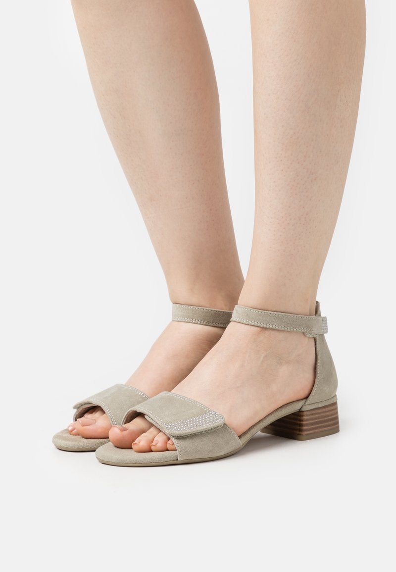 Sandales ouvertes en daim beige avec brides à la cheville et talons carrés bas, portées pieds nus sur un fond blanc.