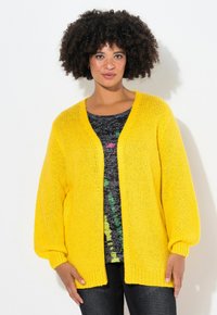 Cardigan giallo a maglia con vestibilità ampia, davanti aperto e polsini a coste. Indossato sopra una maglietta nera a fantasia, con tonalità di verde e rosa.